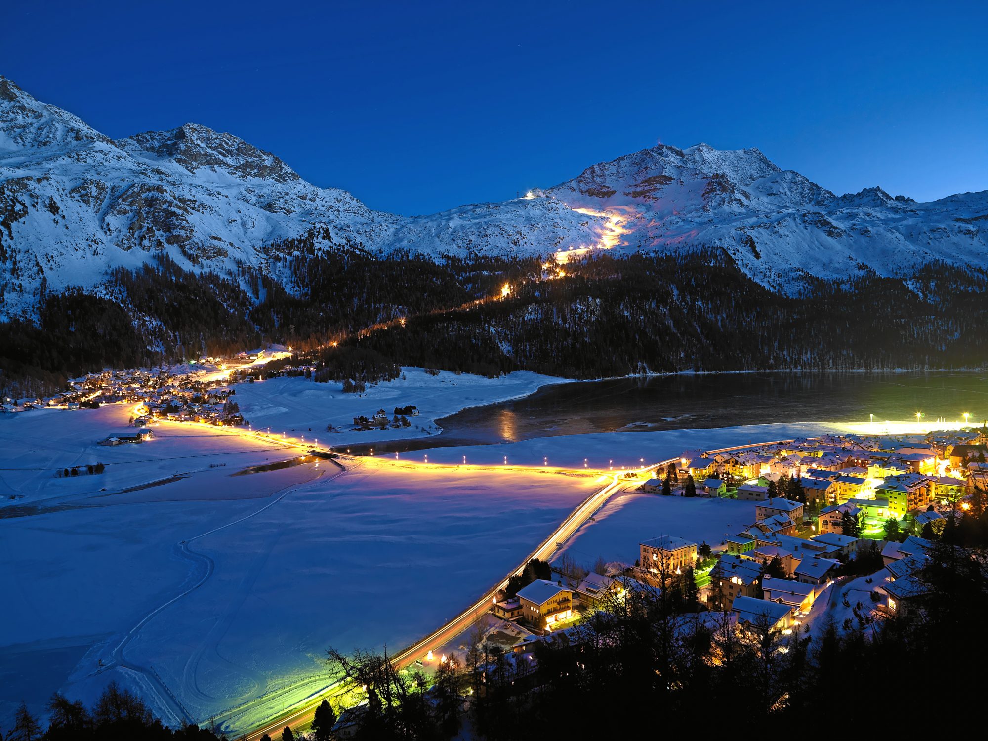 ENGADIN ST. MORITZ - Snow-night at Corvatsch - View from Silvaplana towards the longest illuminated ski slope of Switzerland (4.1 km). The illuminated slope begins at the middle station of Corvatsch and goes along the tree notch of the Surlej slope down to the station at the bottom which is just above the houses of Surlej. In the foreground the lights of the village of Silvaplana. Copyright by ENGADIN St. Moritz By-line:swiss-image.ch/Robert Boesch